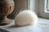Soft white fluffy fur pom on rustic wooden windowsill with vintage wooden decor element in moody natural lighting, creating cozy minimalist composition.