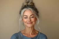 Serene middle-aged woman with silver hair in messy bun smiling peacefully with eyes closed against neutral beige background for wellness and mindfulness content.