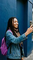 Happy young African American woman with braids taking selfie on smartphone against blue wall wearing denim jacket and purple backpack for social media content.