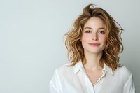 Young Caucasian woman with wavy blonde hair and natural makeup wearing white button-up shirt smiling confidently against light background. Professional headshot style portrait.