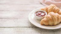 Freshly baked croissants served on white plate with jam and coffee cup on rustic wooden table for breakfast or bakery concepts.