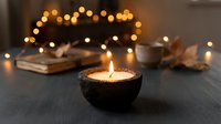 Cozy autumn evening scene with burning candle in dark bowl, warm bokeh lights, old books and dried leaves creating peaceful hygge atmosphere for relaxation.