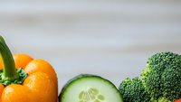 Fresh organic vegetables including orange bell pepper, cucumber slices and green broccoli arranged on white wooden background for healthy cooking and nutrition.