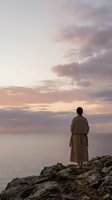 Woman standing on rocky cliff overlooking calm sea at sunset, wearing beige linen dress, contemplating horizon with dramatic pastel sky and clouds, solitude and peaceful reflection concept.