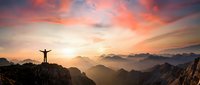 Person with raised arms celebrating success on mountain peak at sunrise with dramatic orange and pink sky over layered mountain ranges creating inspirational moment.