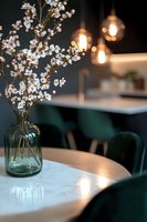 White cherry blossoms in glass vase on marble table with pendant lights creating warm ambiance against dark green restaurant interior background.