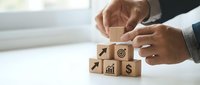 Business professional arranging wooden blocks with financial icons including target, growth chart, and dollar sign on white desk for strategic planning.