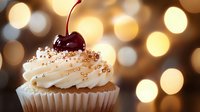 Fresh vanilla cupcake topped with swirled cream frosting, golden sprinkles and dark red cherry against warm bokeh lights background, festive dessert setting.