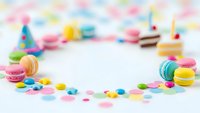 Colorful birthday party setup with macarons, confetti, cake slice and party hats creating festive celebration frame on white background for greeting cards.