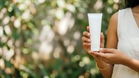 Woman with medium skin tone holding blank white cosmetic tube against blurred green foliage background, suitable for skincare, beauty product marketing, and wellness branding.