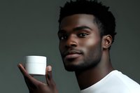 Young African American man in white t-shirt holding white cosmetic jar against dark green background, looking at camera with confident expression for skincare advertising.
