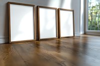 Three wooden frame mockups standing on hardwood floor against white wall near window, blank canvas ready for artwork presentation in modern interior.
