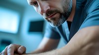 Close up portrait of mature bearded man in blue t-shirt looking down thoughtfully while working or studying in modern office environment with soft lighting.