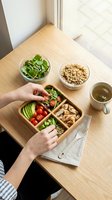 Overhead view of woman's hands holding wooden meal prep box with fresh vegetables, grains, and protein on light table near window for healthy eating and nutrition planning.