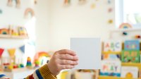 Child's hand holding blank white card in colorful playroom with toys, educational materials, and decorations for greeting, announcement, or creative project messaging.