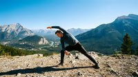 Young Caucasian man practicing yoga warrior pose on rocky mountain summit with stunning valley and peaks under clear blue sky.