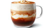 Layered caramel macchiato coffee drink in clear glass mug showing distinct cream, milk and espresso layers topped with whipped cream and caramel drizzle on white background.