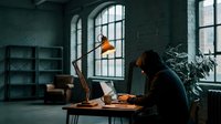 Young woman with dark hair, wearing black clothing, sitting at wooden desk working on laptop in industrial loft space with large windows, brick walls, shelving, and warm pendant lighting for creative workspace.