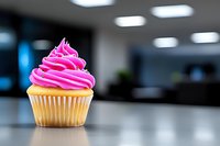 Vibrant pink frosted vanilla cupcake on reflective surface against blurred modern interior background, fresh baked dessert with swirled buttercream topping.