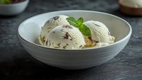 Creamy vanilla ice cream scoops with chocolate chips in white ceramic bowl garnished with fresh mint leaves on dark slate surface for dessert menu.