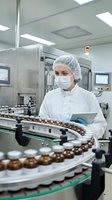 Female pharmaceutical technician in sterile cleanroom monitoring automated production line with tablet computer for quality control and manufacturing processes.
