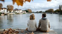 Couple in cozy sweaters sitting by riverside in autumn, enjoying peaceful view of water and distant houses under cloudy sky with falling leaves.