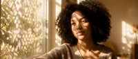 Young African American woman with curly hair smiling warmly in golden sunlight streaming through window, creating peaceful morning atmosphere for lifestyle content.