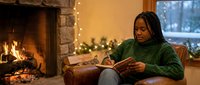 Young African American woman in green sweater reading book by cozy fireplace with Christmas lights creating warm holiday atmosphere at home.