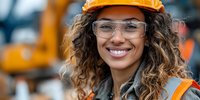 Smiling mixed-race female construction worker wearing orange hard hat and safety glasses at building site with heavy machinery in background.
