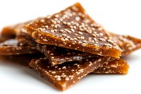 Crunchy toffee brittle pieces with white sesame seeds scattered on top, stacked in layers showing rich caramelized texture and golden brown color on white background.