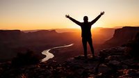 Silhouette of person with raised arms celebrating success on mountain cliff overlooking canyon river at golden sunrise for motivation and achievement concepts.