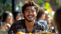 Young Asian man with bright genuine smile at outdoor restaurant gathering, wearing patterned shirt, enjoying social moment with friends during daytime celebration.