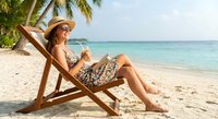 Young woman relaxing on wooden beach chair under palm tree, enjoying tropical vacation with drink. Summer holiday leisure concept for travel marketing.