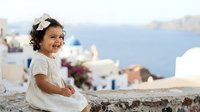 Young girl with curly dark hair wearing white dress and flower crown sitting on stone wall overlooking coastal village with white buildings and blue sea, suitable for travel and family lifestyle content.