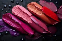 Smeared lipstick swatches in vibrant shades of pink, red, and purple on dark background with water drops, showing rich creamy texture and pigmentation.