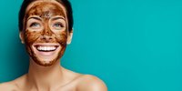 Woman with chocolate facial mask smiling against turquoise background, showcasing skincare routine for beauty and wellness treatments.