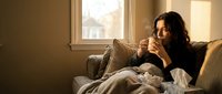 Woman with long dark hair, South Asian ethnicity, sitting on a beige armchair by a window holding a warm beverage in cozy home setting, relaxed lifestyle moment for wellness and comfort themes.