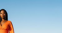 Woman with long braided hair wearing bright orange wrap top against clear blue sky, suitable for lifestyle and fashion marketing campaigns.