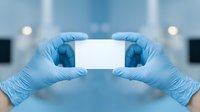 Healthcare professional wearing sterile blue latex gloves holding blank white card in laboratory or medical facility setting for clinical communication and branding purposes.