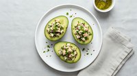 Halved avocados topped with quinoa, feta cheese, and fresh herbs served on white plate for healthy lunch or vegetarian meal preparation.