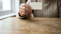 Senior man with weathered hands holding blank white card at wooden table, suitable for business cards, invitations, promotions, or marketing campaigns.