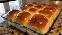 Freshly baked golden dinner rolls in glass baking dish on granite countertop, showing soft texture and glossy buttered tops with professional lighting.