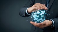 Businessman in dark suit holding glowing skill up icon with gear symbol and upward arrow, representing professional development, training, and career advancement in business.
