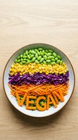Colorful vegan bowl with edamame, corn, red cabbage, and shredded carrots spelling VEGAN, shot from above on a wooden surface.