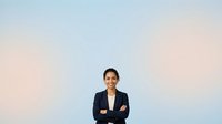 Young woman with dark hair and glasses, South Asian ethnicity, wearing a navy blazer and white shirt, standing with crossed arms against a soft gradient background transitioning from blue to pink, suitable for corporate, business, and professional website use.