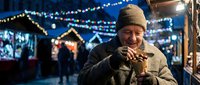 Elderly Caucasian man in warm winter clothing enjoying hot beverage at festive Christmas market with twinkling lights and holiday decorations in background.