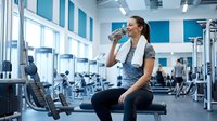 Young athletic woman drinking water from bottle during workout break in modern fitness center with exercise equipment and large windows.