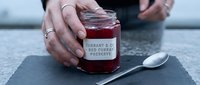 Person holding jar of red currant jam with spoon on slate surface, homemade preserve in glass container with label for food styling and culinary content.