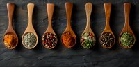 Aromatic spices and herbs in wooden spoons arranged in row on dark stone background, colorful seasoning mix including turmeric, oregano, peppercorns and parsley.