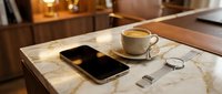Smartphone, analog watch, and espresso coffee cup on a marble counter in a warm, elegant cafe interior setting.
